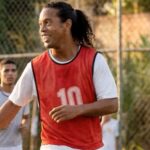 Ronaldinho Gaúcho em campanha publicitária segurando cerveja em cenário descontraído que remete a campo de futebol