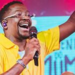 Cantor Mumuzinho se apresenta no palco durante show da turnê Resenha do Mumu, com público e estrutura de evento musical ao fundo