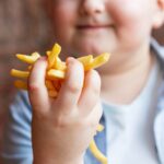 Criança segurando alimentos industrializados em contraste com frutas, em alerta para obesidade infantil causada por ultraprocessados.