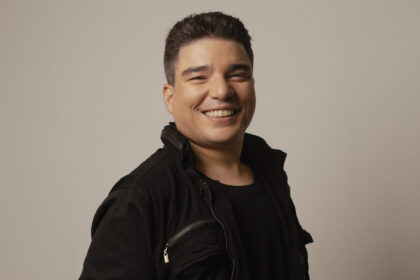 Arthur Espíndola posa sorrindo para fotografia em estúdio, usando roupa preta diante de fundo neutro.