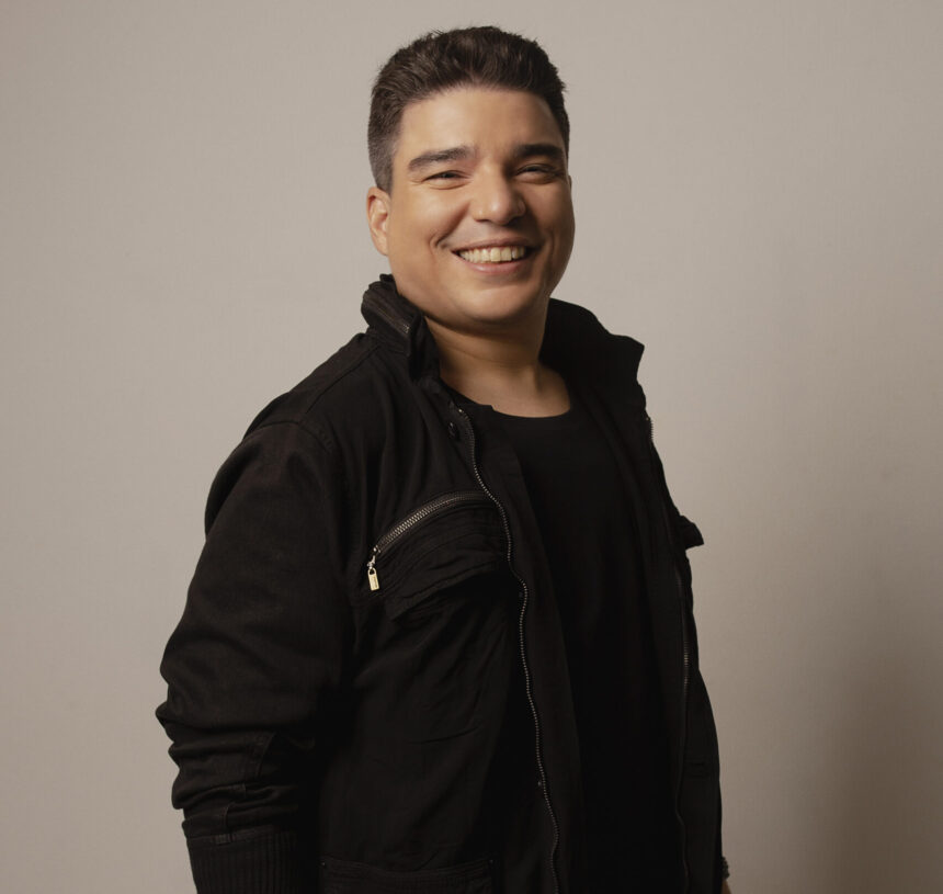 Arthur Espíndola posa sorrindo para fotografia em estúdio, usando roupa preta diante de fundo neutro.