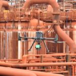 Drone industrial realizando inspeção técnica em área operacional de refinaria com estruturas metálicas e equipamentos industriais ao fundo.