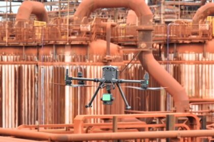Drone industrial realizando inspeção técnica em área operacional de refinaria com estruturas metálicas e equipamentos industriais ao fundo.