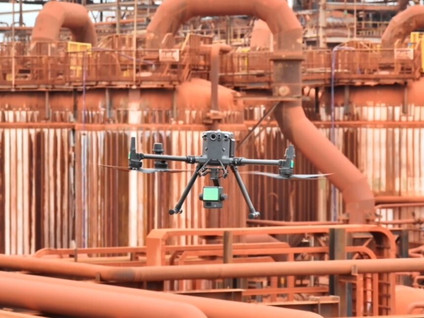 Drone industrial realizando inspeção técnica em área operacional de refinaria com estruturas metálicas e equipamentos industriais ao fundo.