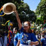 Brincante do Arrastão do Pavulagem ergue chapéu decorado com fitas coloridas durante cortejo cultural nas ruas de Belém, cercada por participantes vestidos de azul em meio à celebração popular ao ar livre.