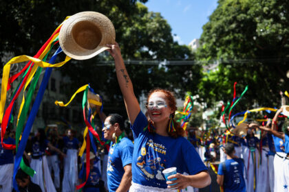 Brincante do Arrastão do Pavulagem ergue chapéu decorado com fitas coloridas durante cortejo cultural nas ruas de Belém, cercada por participantes vestidos de azul em meio à celebração popular ao ar livre.