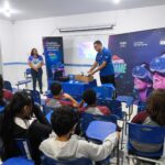 Alunos sentados em sala de aula acompanham apresentação sobre tecnologia e sustentabilidade enquanto educadores demonstram equipamentos de realidade virtual em escola pública.