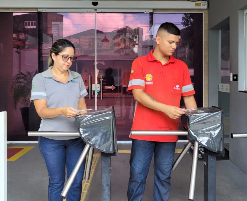Mãe e filho passam juntos pela catraca de acesso de uma fábrica em Belém, usando uniforme da empresa durante a rotina de trabalho.