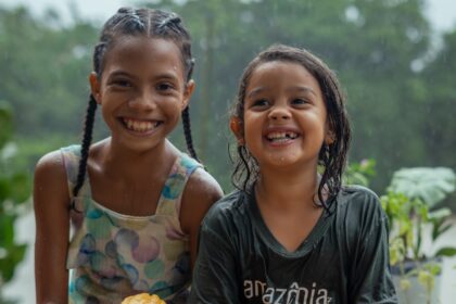 Duas crianças ribeirinhas sorriem sentadas em um trapiche de madeira em Barcarena, no Pará, cercadas por frutos de cacau durante chuva em cenário amazônico.