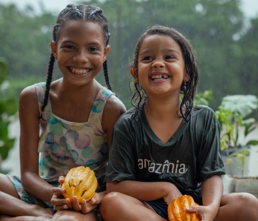 Duas crianças ribeirinhas sorriem sentadas em um trapiche de madeira em Barcarena, no Pará, cercadas por frutos de cacau durante chuva em cenário amazônico.